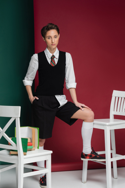 stylish young woman with short hair posing in school uniform around chairs and books on green and pink background  - Foto, Imagen