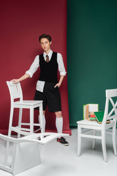 full length of young woman with short hair posing in school uniform around chairs and books on green and pink background  - Foto, immagini