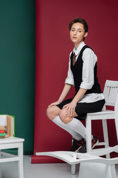 pełna długość stylowy młody student z krótkimi włosami siedzi na krześle w pobliżu książek na zielonym i czerwonym tle  - Zdjęcie, obraz