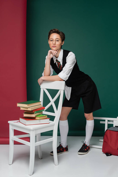 täyspitkä tyylikäs nuori nainen lyhyet hiukset nojaten tuoli kirjoja vihreä ja vaaleanpunainen tausta  - Valokuva, kuva