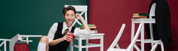 felice giovane studente in occhiali che tiene bevanda in tazza di plastica vicino a sedie e libri su sfondo verde e bordeaux, banner  - Foto, immagini