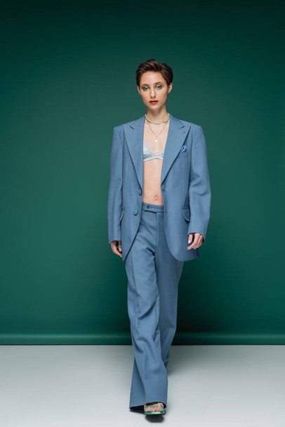 full length of young woman with short hair walking in blue pantsuit on turquoise green background  - Фото, зображення