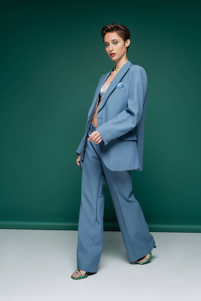 full length of young woman with short hair walking in blue pantsuit with bra on turquoise green background  - Φωτογραφία, εικόνα