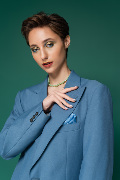 young woman with short hair posing in blue blazer and looking at camera on turquoise green background  - Photo, image
