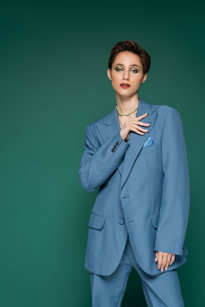 elegant young woman with short hair posing in blue blazer and looking at camera on turquoise green background  - Foto, afbeelding