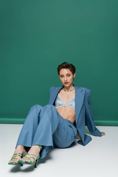 full length of young woman with short hair posing in blue suit with satin bra and heeled sandals on turquoise green  - Foto, imagen