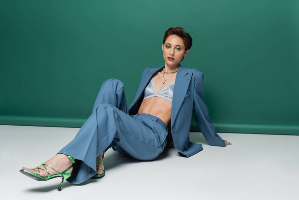 full length of young woman with short hair posing in stylish suit with satin bra and heeled sandals on turquoise green  - Photo, image