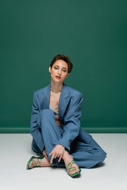 full length of young woman with short hair posing in stylish suit and heeled sandals on turquoise white background  - Fotó, kép