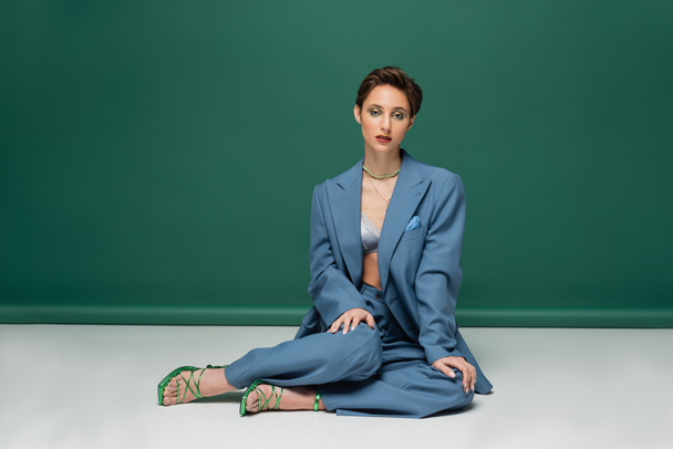 full length of young woman with short hair posing in trendy suit and heeled sandals on turquoise white background  - Zdjęcie, obraz