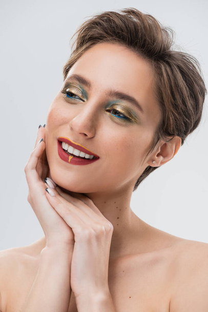 glückliche junge Frau mit kurzen Haaren und hellem Make-up, die isoliert von grau wegschaut  - Foto, Bild
