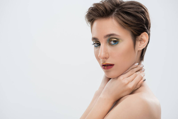 portrait of young woman with bright makeup touching neck and looking at camera isolated on grey - Photo, image