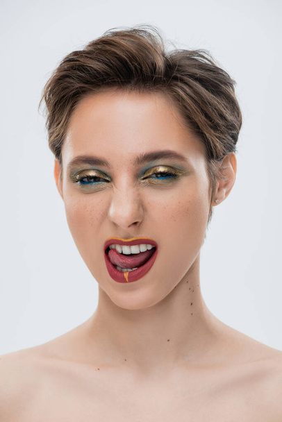 portrét emocionální mladé ženy s lesklým make-up a krátké vlasy vyčnívající jazyk izolované na šedé  - Fotografie, Obrázek