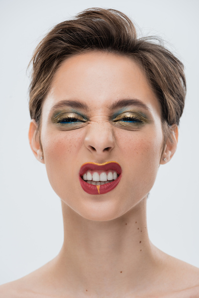 portret van emotionele jonge vrouw met glanzende make-up en kort haar grijnzend geïsoleerd op grijs  - Foto, afbeelding