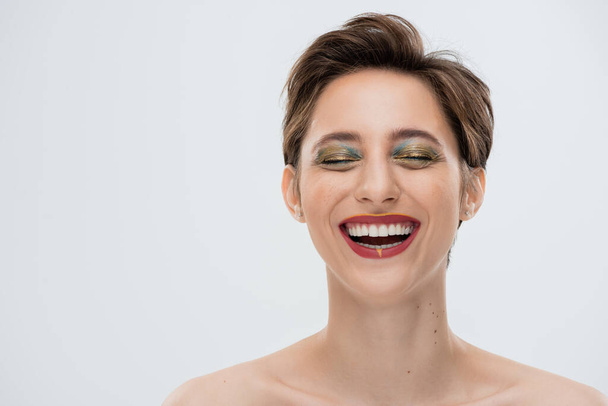 positive young woman with shiny makeup and short hair smiling isolated on grey  - Фото, изображение