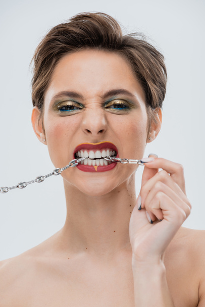retrato de mulher jovem com maquiagem brilhante e cabelo curto mordendo cadeia de prata isolado em cinza  - Foto, Imagem