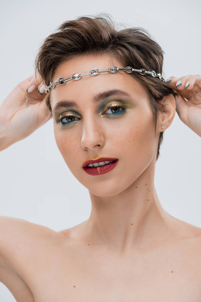 young woman with shiny makeup and short hair wearing silver chain on head isolated on grey  - Zdjęcie, obraz