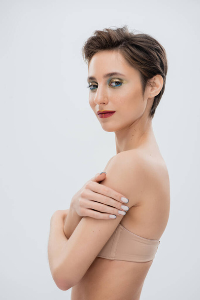pretty young woman with shimmery eye makeup standing in strapless bra isolated on grey  - Foto, Imagem