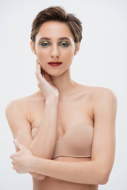 hübsche junge Frau mit glänzenden Augen Make-up steht in trägerlosem BH isoliert auf grau  - Foto, Bild
