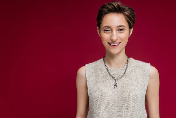 pleased young woman in sleeveless shirt and chain necklace standing on maroon background  - Photo, Image