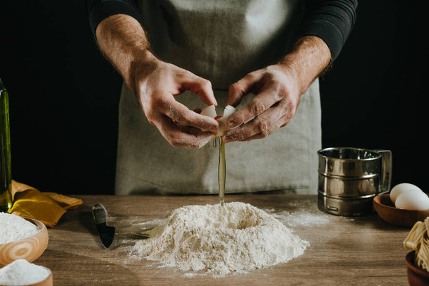 Unrecognizable man kneading dough on wooden background against dark wall. Close-up of male hands cutting egg. Stage 2 of preparation homemade noodles. Selective focus. - Fotó, kép