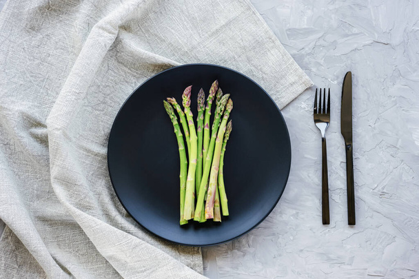Fresh green asparagus on black plate on grey concrete background, cutlery and napkin. Healthy vegan raw food concept. Top view, flat lay. - Фото, изображение