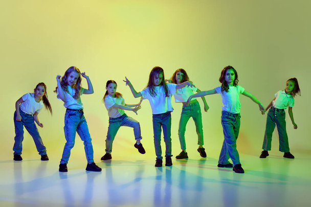 Movements. Little cute girls, children in casual clothes dancing, learning hip-hop style against green studio background in neon light. Concept of childhood, hobby, sportive lifestyle - Фото, зображення