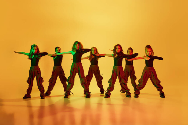 Group of little girls, children, talented dancers in stylish sportive clothes dancing against yellow studio background in neon light. Concept of childhood, hobby, sportive lifestyle, dance class - Photo, image