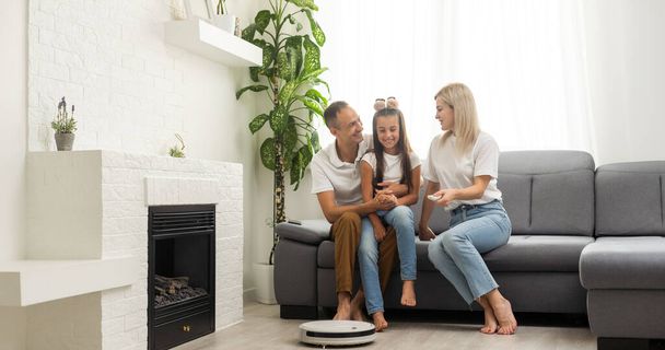 Young family resting on the couch while robotic vacuum cleaner doing its work at home. Household robots concept - 写真・画像