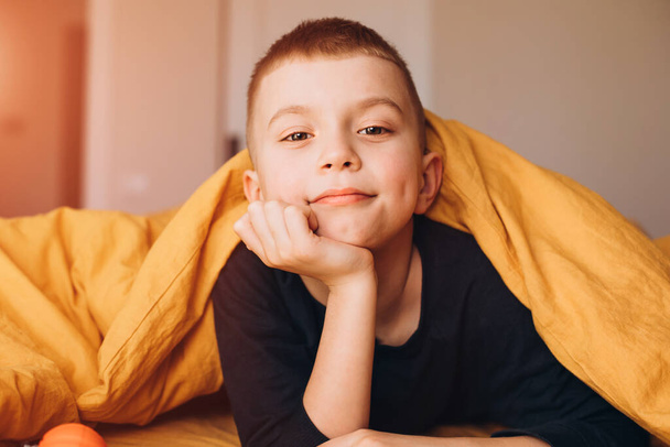 A joyful boy peers out from under the yellow blanket. The boy in his bad. Front view - Foto, imagen