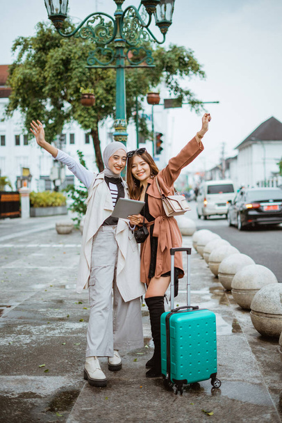 two excited travellers raising their hands while standing together at the sidewalk bring their travel equipment - Photo, Image