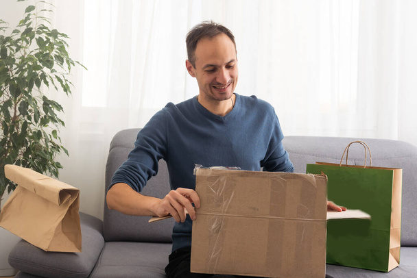 Подарунок від далекого друга. Щасливий тисячолітній чоловік розпаковує відкриту коробку з подарунковим сюрпризом на день народження, отриманим поштою. Усміхнений молодий хлопець із задоволенням отримує пакет із споживчими товарами, придбаними онлайн
 - Фото, зображення