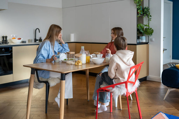 Samotna matka z dziećmi siedzi przy stole kuchennym i je śniadanie przed szkołą. Rutyna rodzinna. Nastoletnia córka rozmawia z mamą i młodszym bratem o planie studiów. Zdrowe jedzenie razem - Zdjęcie, obraz