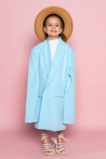 Pienen tytön muotokuva ylisuuri aikuinen sininen bleiseri ja pyöreä ruskea hattu. Lapsella on aikuisten vaatteet. Eristetty vaaleanpunaisella pohjalla. - Valokuva, kuva