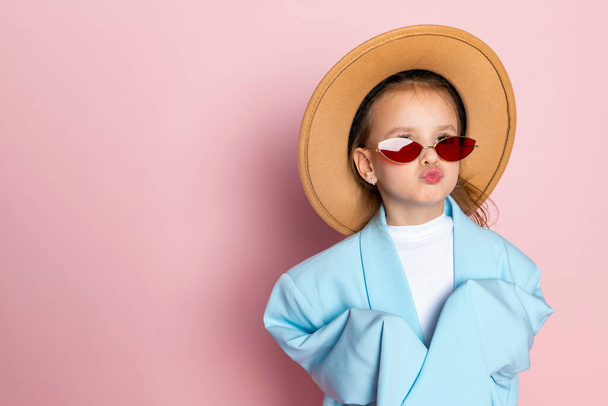 Портрет маленької дівчинки в синьому блакитному блістері для дорослих, стильні червоні окуляри і круглий коричневий капелюх. Дитина носить дорослий одяг. Ізольовані на рожевому фоні
. - Фото, зображення