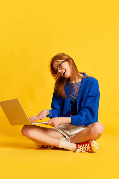 Amistad en línea, comunicación. Una chica alegre, estudiante, adolescente con gafas que sostienen el ordenador portátil y sonriendo en el fondo del estudio amarillo. Redes sociales, amigos, jóvenes, concepto de relación - Foto, imagen
