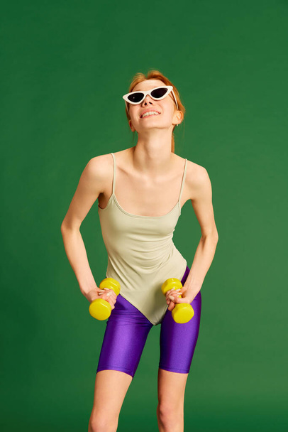 Retro style workout. Sporty red haired young girl wearing vintage sportswear training with dumbbells on green studio background. Sport, fitness, healthy life style, fashion, activity, youth concept - Φωτογραφία, εικόνα