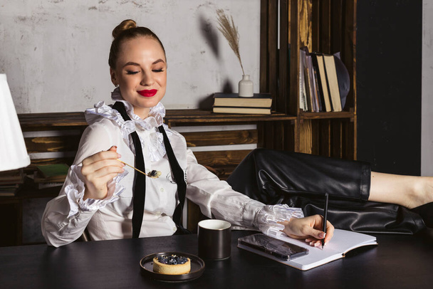 Piękna kobieta w eleganckiej koszuli vintage i jasnoczerwonej szmince siedząca przy czarnym drewnianym biurku, pracująca i jedząca małe ciasto jagodowe. - Zdjęcie, obraz