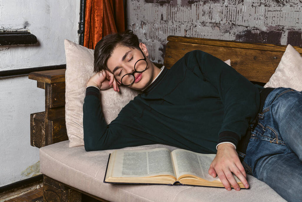 Um estudante diligente, bem vestido de óculos redondos, adormeceu em um sofá com um livro em suas mãos. O estudante adormeceu enquanto estudava. - Foto, Imagem
