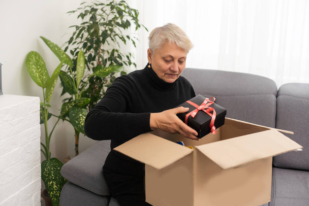 Sonriente cliente anciana recibir paquete de envío posterior en casa, feliz anciana abuela de edad mantenga abierta caja de cartón sentarse en el sofá en la sala de estar, pedido de compras en línea concepto de entrega de mensajero rápido
 - Foto, Imagen