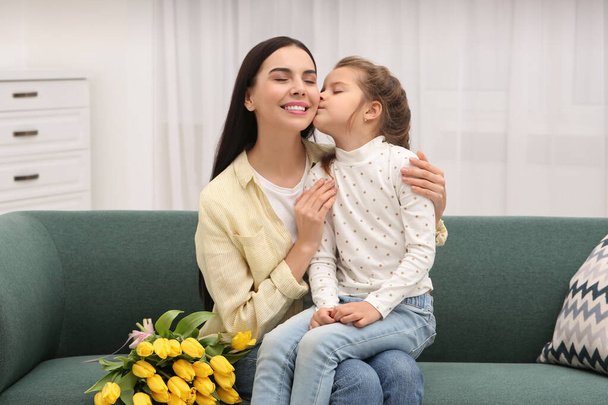 Happy woman and her cute daughter with bouquet of yellow tulips on sofa at home. Mother's day celebration - Fotoğraf, Görsel
