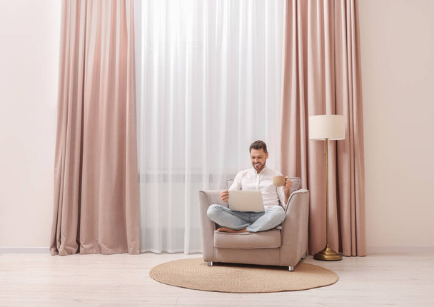 Smiling man drinking tea while working on laptop near window with beautiful curtains at home. Space for text - Photo, Image