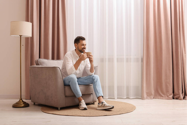 Smiling man drinking tea while resting on armchair near window with beautiful curtains at home. Space for text - Foto, Bild