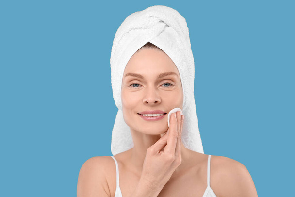Beautiful woman in terry towel removing makeup with cotton pad on light blue background - Φωτογραφία, εικόνα
