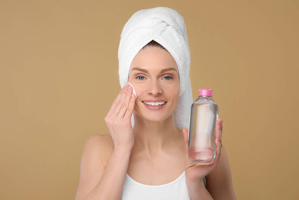 Beautiful woman in terry towel removing makeup with cotton pad on beige background - Foto, Imagen