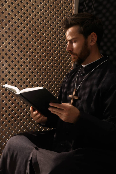 Catholic priest in cassock reading Bible in confessional booth - Photo, image