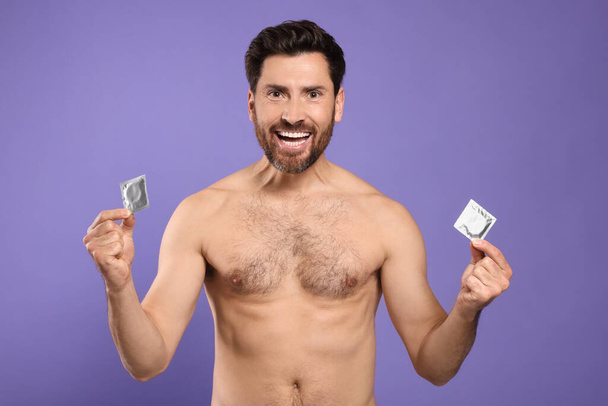 Emotional naked man holding condoms on purple background. Safe sex - Photo, image