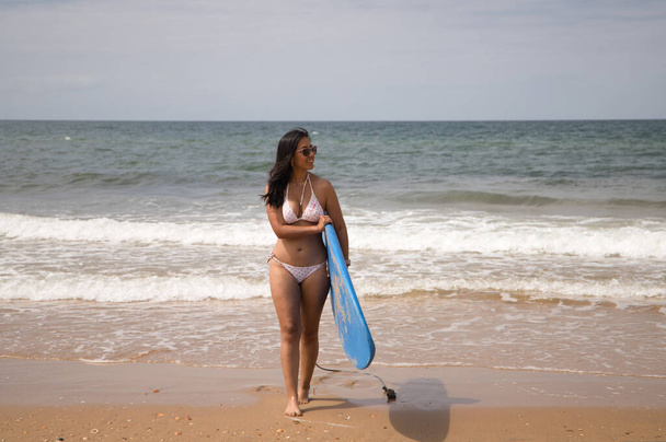 Schöne junge lateinamerikanische Frau im Bikini, die mit blauem Surfbrett aus dem Meer kommt. Die Frau liegt am Strand. Ferien- und Sommerkonzept. - Foto, Bild