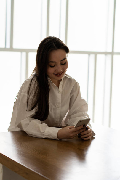 Μια νεαρή μελαχρινή κάθεται σε ένα τραπέζι σε ένα φωτεινό δωμάτιο με ένα τηλέφωνο στα χέρια της. Μια μελαχρινή με λευκό πουκάμισο συνομιλεί με φίλους μέσω αγγελιοφόρου. Υψηλής ποιότητας φωτογραφία - Φωτογραφία, εικόνα