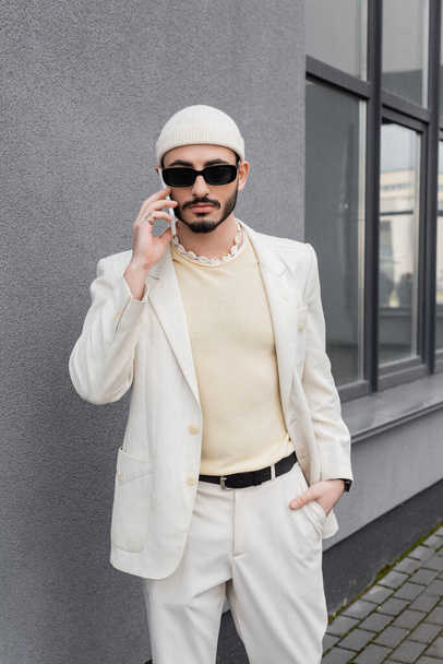 Trendy gay man in sunglasses and beige suit talking on smartphone outdoors - 写真・画像