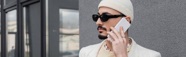 Trendy gay man in sunglasses and hat talking on cellphone near building outdoors, banner  - Fotografie, Obrázek
